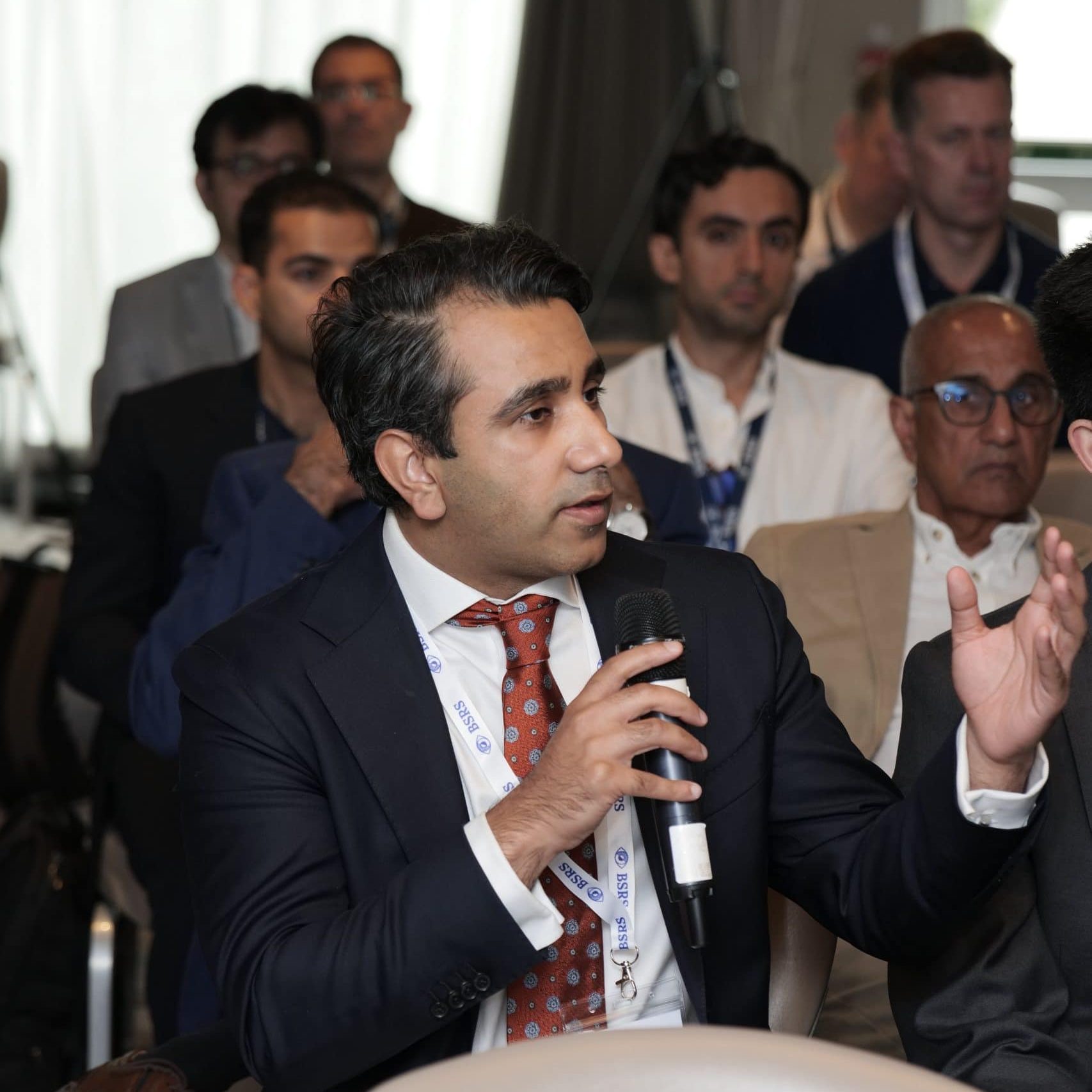 A male delegate in a suit speaks into a microphone during a session at the BSRS conference, with other attendees seated and listening in the background.